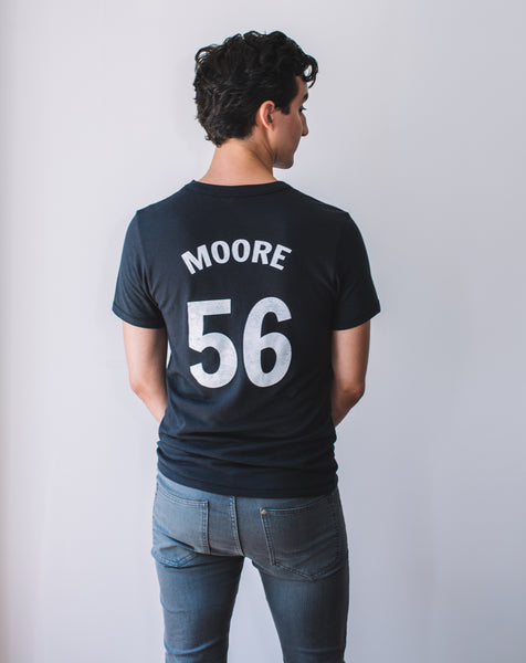 Back of Latinx male model wearing a Marianne Moore #56 black unisex baseball tee